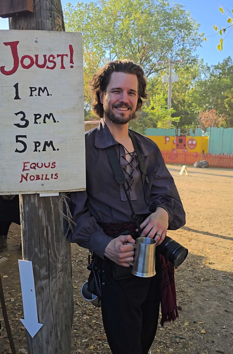 Man in pirate costume holding a tankard and camera next to a Joust! sign at a fair