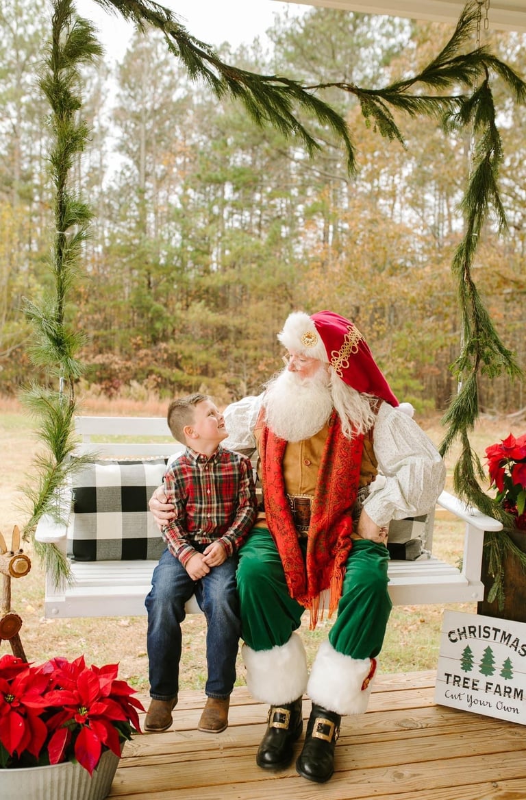 A young boy sits with Santa Claus on a white porch swing at a Christmas tree farm.