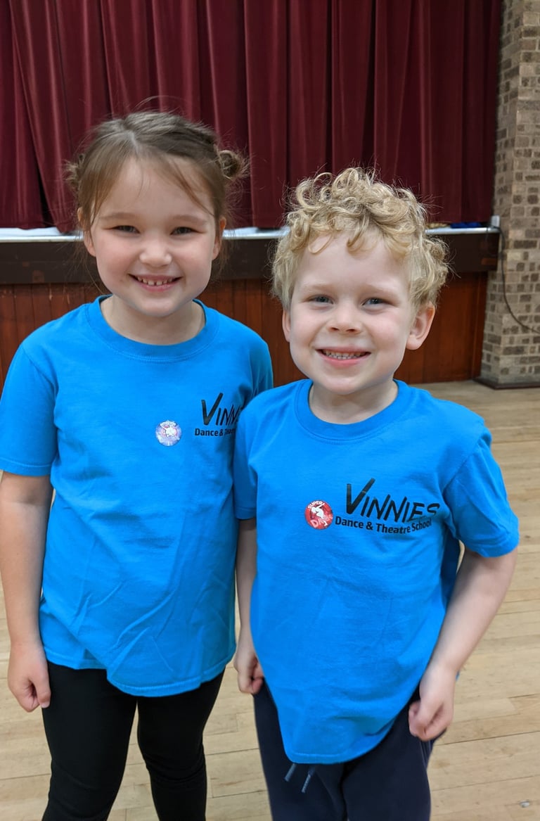 Two smiling children in blue Vinnies Dance & Theatre School t-shirts on a wooden dance floor.