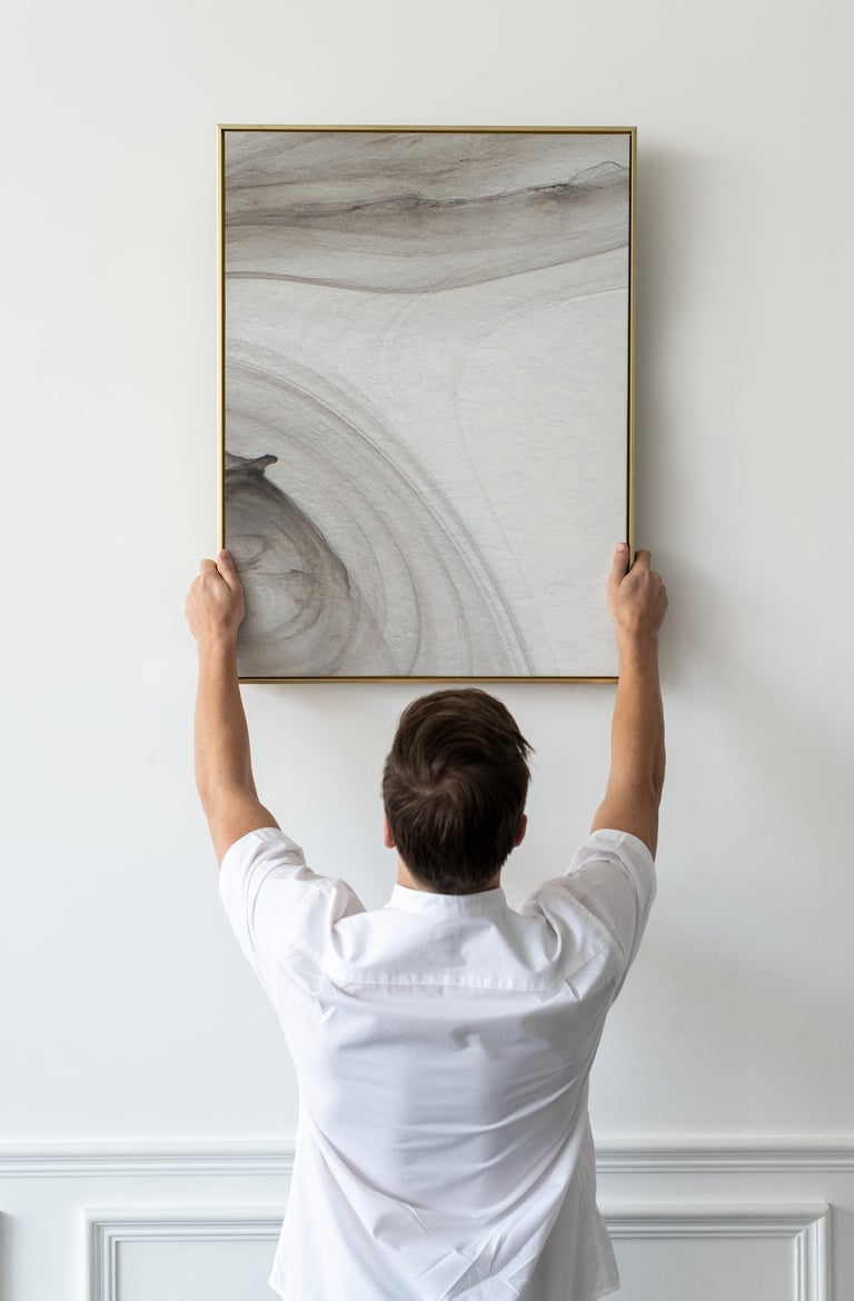 A man hanging a large abstract neutral-toned canvas painting in a gold frame on a white wall.