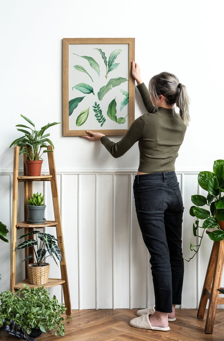 A woman hanging a wooden framed botanical leaf art print on a white wall in a room filled with indoor plants.