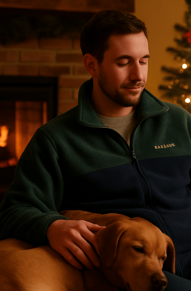 Man wearing Columbia fleece jacket sitting by fireplace with dog beside him and Christmas tree