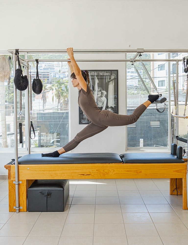 Woman doing a split stretch on a Pilates Cadillac apparatus in a bright studio in Cabo San Lucas.
