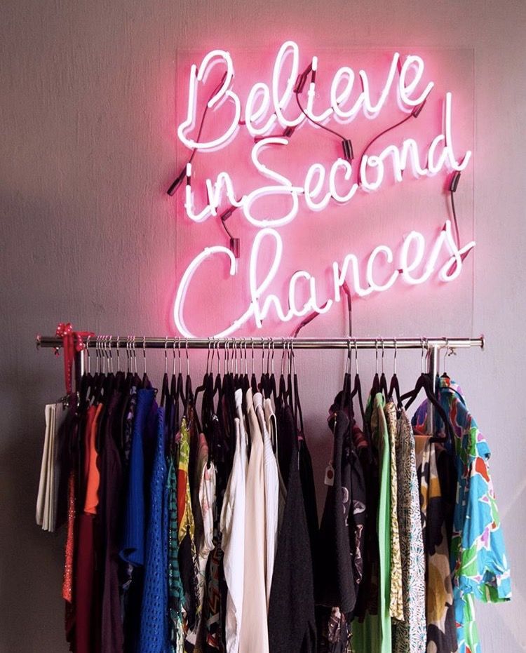 Letrero de neón rosa que dice “Cree en segundas oportunidades” sobre rack de ropa de segunda mano.