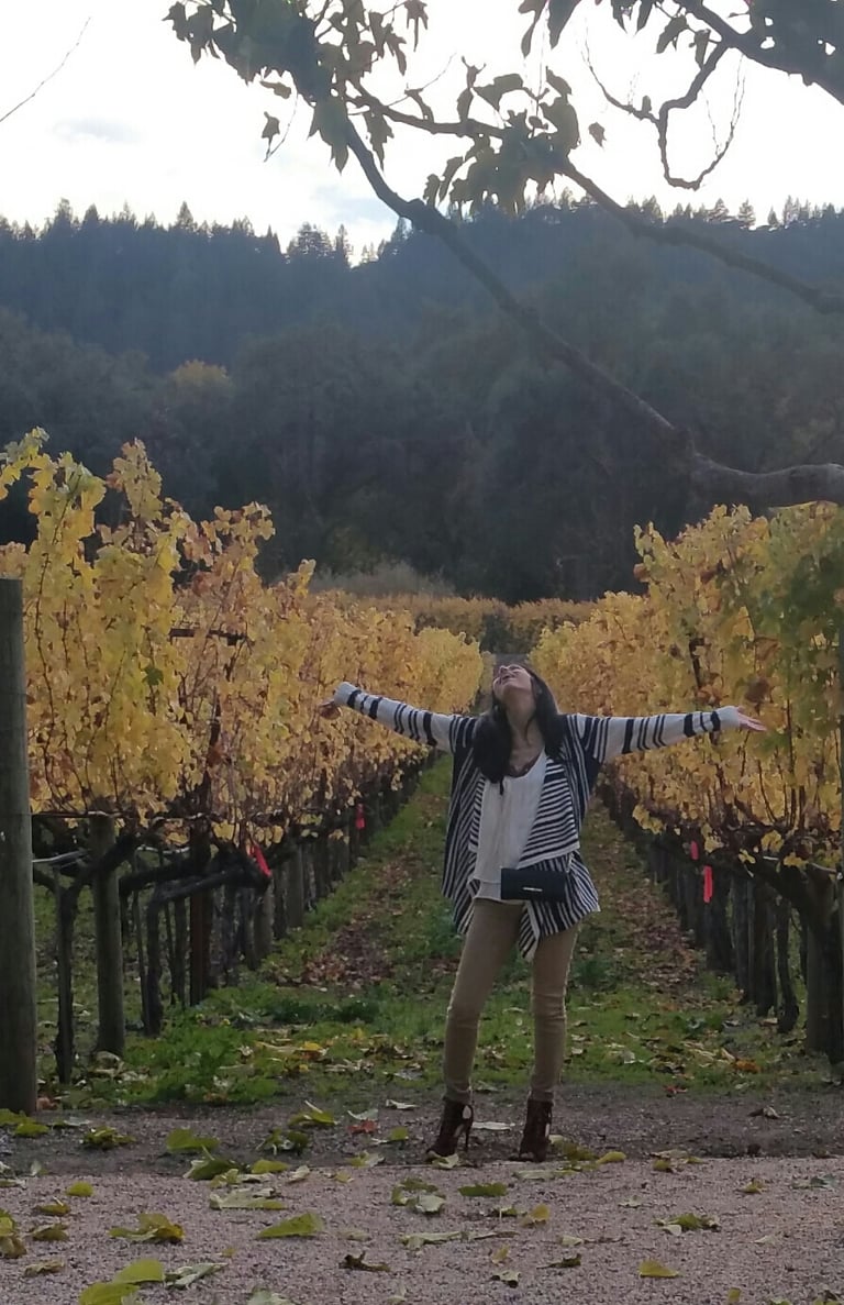 Photograph of Rebecca with hands in the air and a scenic vineyard background.