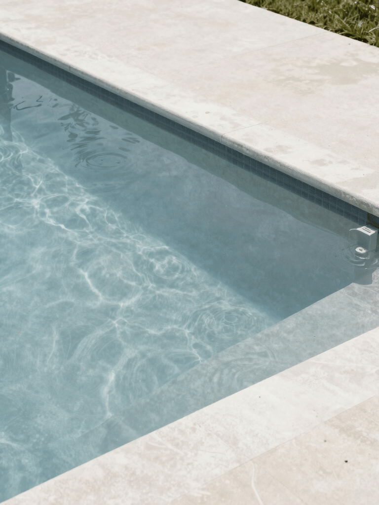 A high-angle shot of a classic North American suburban pool at midday. The water is a clear, soft blue-grey, contrasting with a clean off-white concrete deck. Minimalist, contemporary aesthetic.