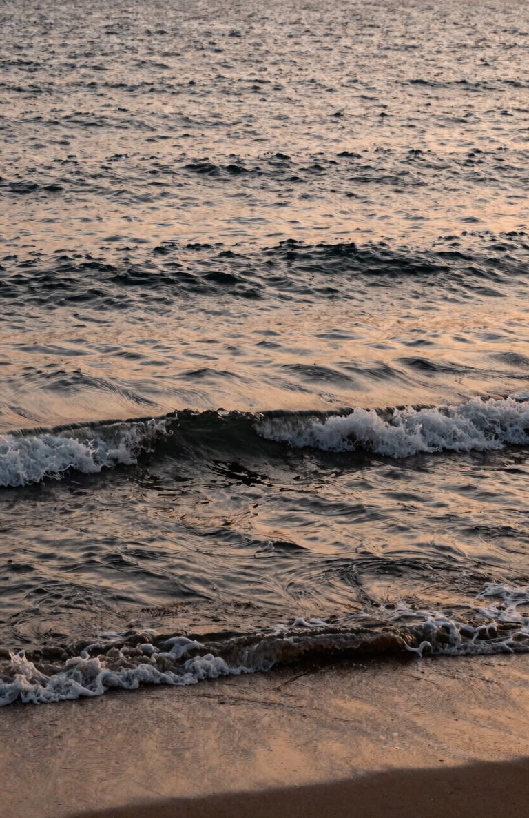 A serene photograph of the Mediterranean sea at sunset, with waves gently rolling onto a sandy shore, emphasizing the organic warm beige and ebony tones of the water.