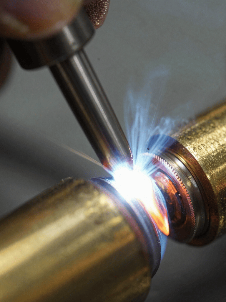 Macro photography of a perfectly executed TIG weld on a brass joint. The metal shows a soft, muted bronze hue with technical precision in the bead. Shallow depth of field with a dark slate grey background.