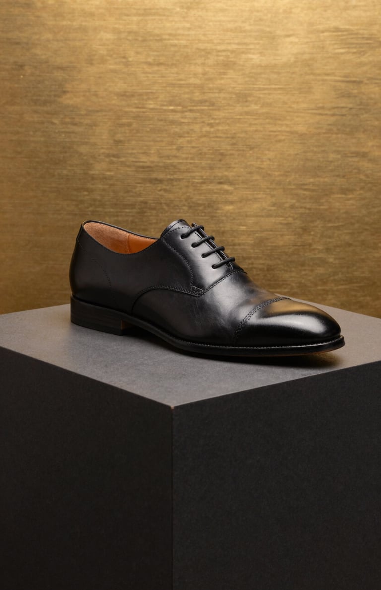 High-end product photography of a luxury leather shoe on a dark charcoal black pedestal. The background is brushed gold, with sharp lighting and clean lines. Modern and commercial style.