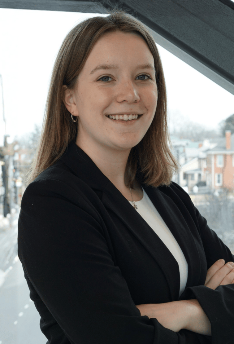 A smiling young professional woman in a black blazer and white top with arms crossed.