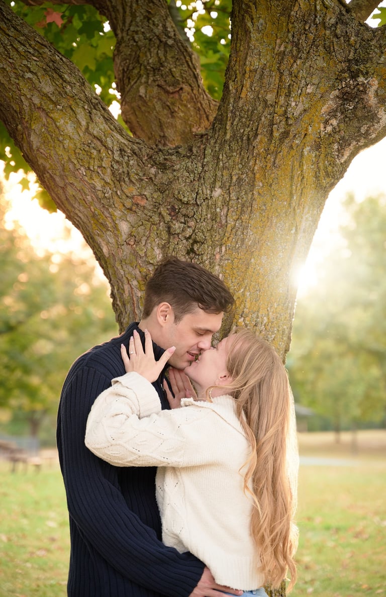 a man and woman kissing in front of a tree with sunset behind in a park