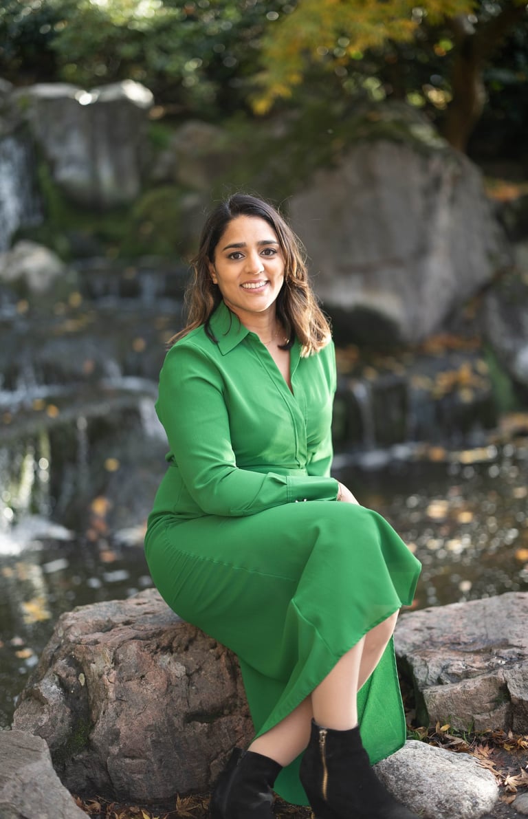 A photo of Anita wearing a green dress. She is sat on a rock with a water feature in the background.