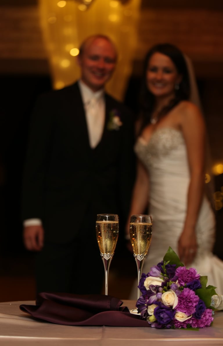 Champagne Glasses and Bridal Bouquet on table in focus. Bride and Groom Looking at camera smiling.