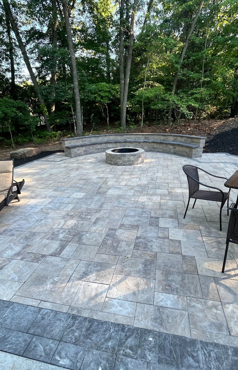 Custom stone paver patio featuring a round fire pit, curved seating wall, lounge chairs, and a dining set.