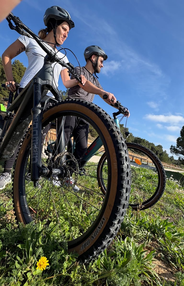 Two mountain bikers in helmets standing on a grassy trail with their mountain bikes on a sunny day.