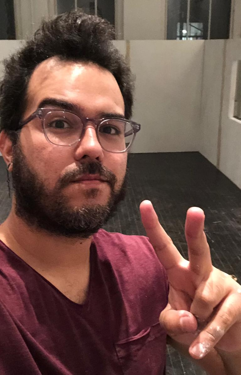 A man with glasses and a beard making a peace sign in an art studio with white partitions.