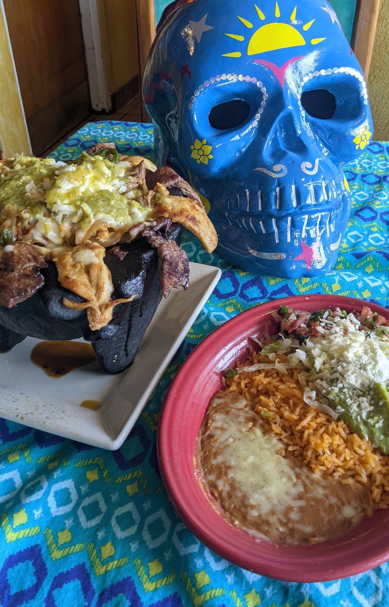 Traditional Mexican molcajete with grilled meats next to rice, beans, and a blue decorative sugar skull.