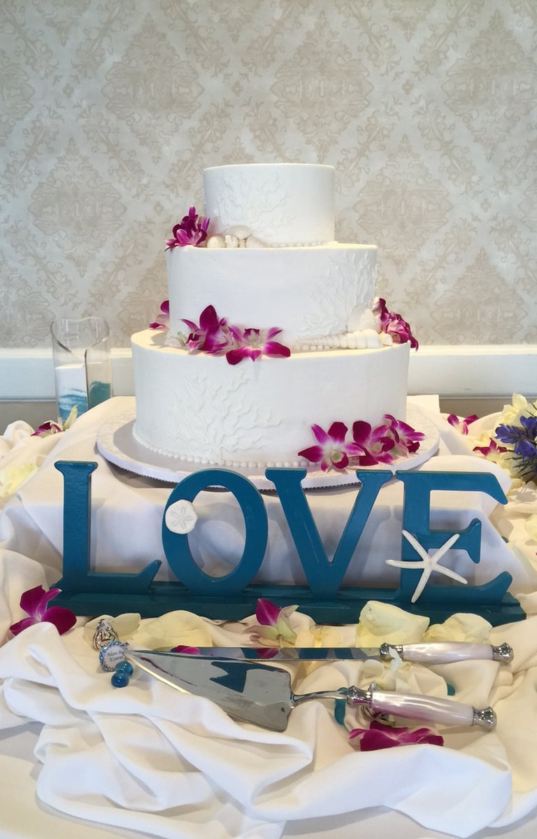 three-tier white wedding cake decorated with fuchsia orchids and a teal“love” sign in front, 