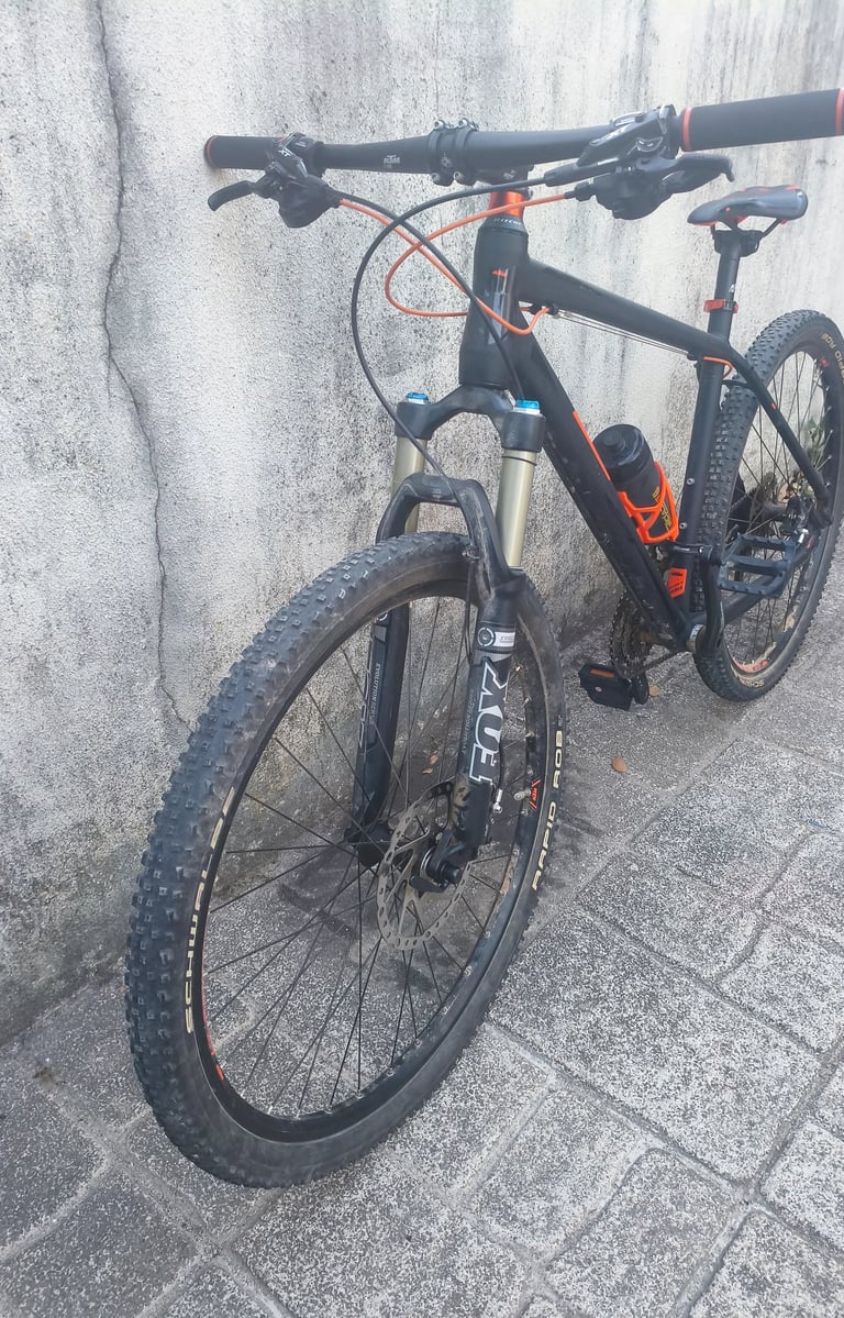 Bicicleta de montanha KTM preta e laranja com suspensão dianteira em destaque, vista frontal em três