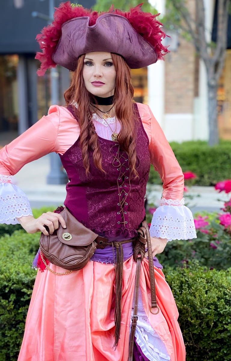 A woman in a detailed pirate costume with a feathered tricorn hat and corset standing in a garden.
