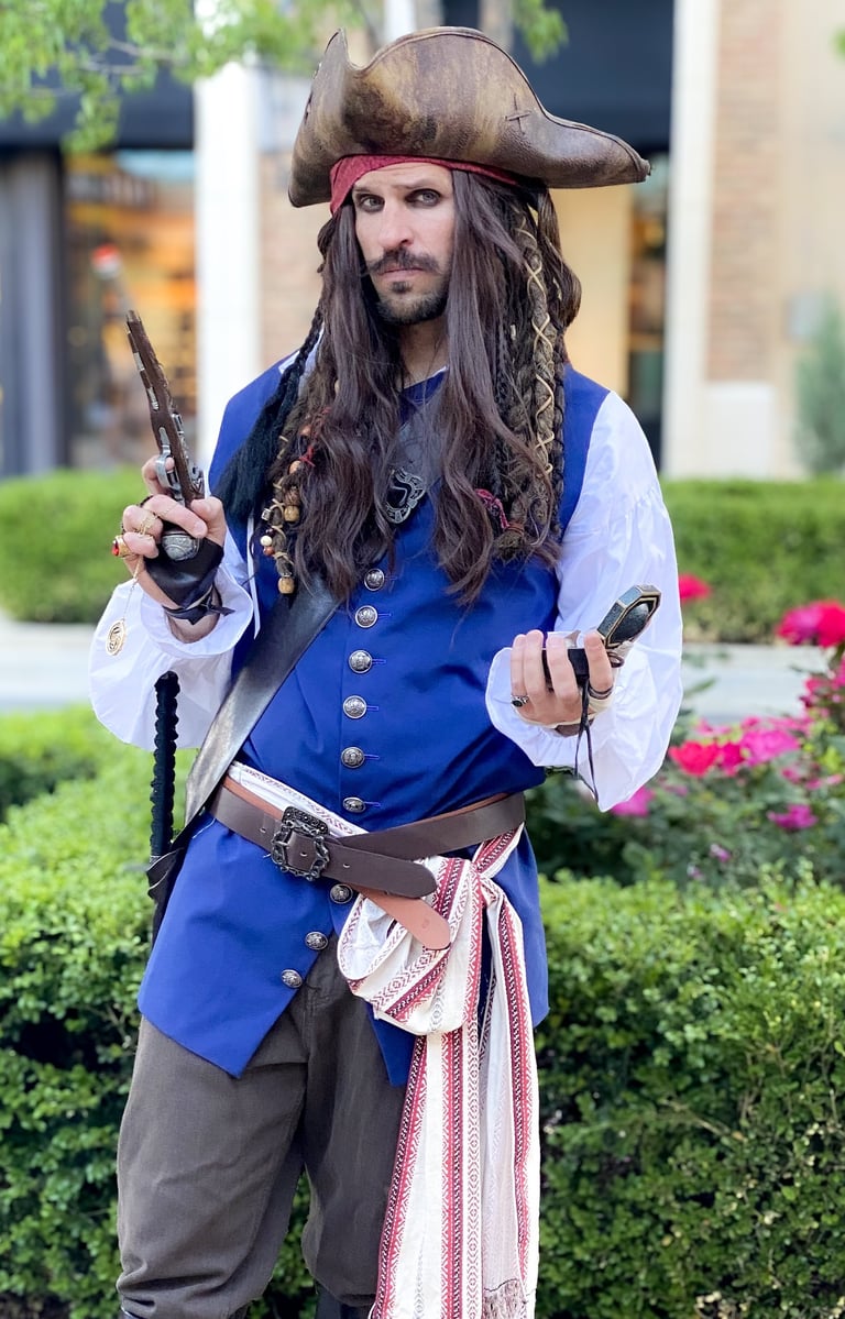 A man in a detailed Jack Sparrow pirate costume holding a flintlock pistol and compass in a garden.