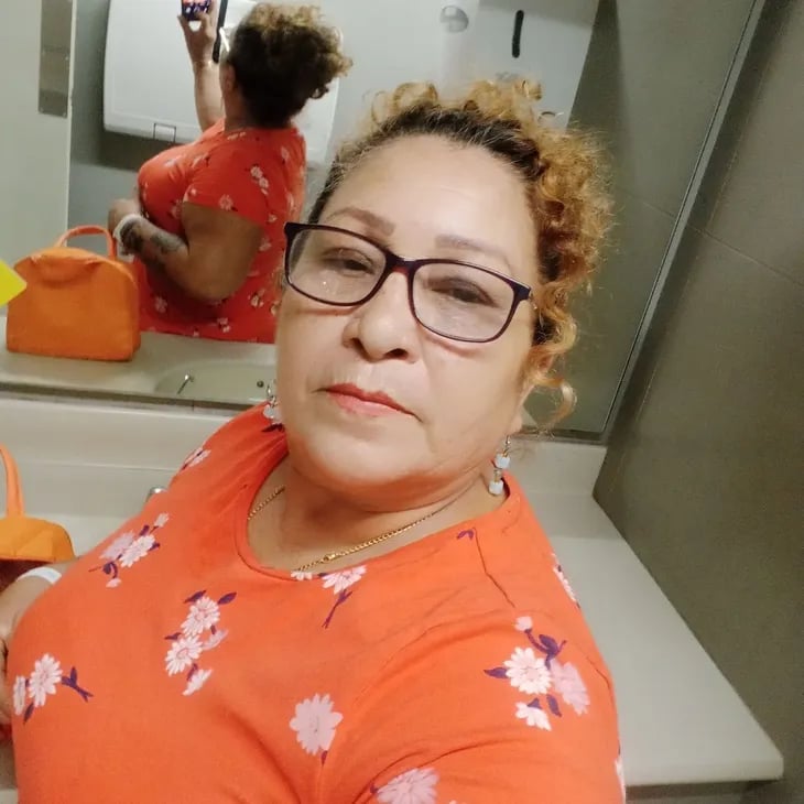 A woman with curly hair and glasses taking a mirror selfie in an orange floral top.