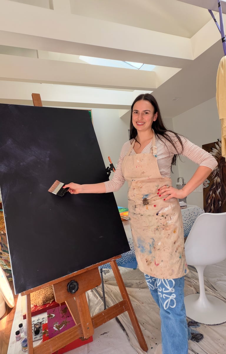 artist in a paint-stained apron holds a brush against a large black canvas in her studio.