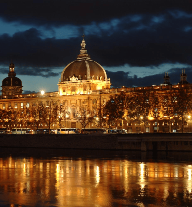 Vue nocturne de l'Hôtel-Dieu de Lyon illuminé, avec ses lumières dorées se reflétant dans le Rhône.