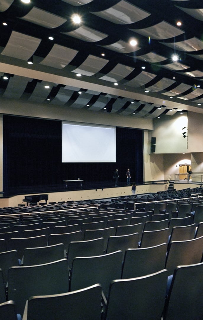 Large empty auditorium with rows of chairs facing a stage and projection screen, under a ceiling wit