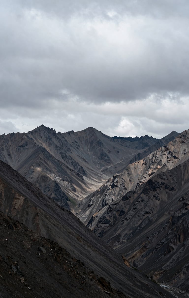A wide shot of a jagged mountain range under a heavy, overcast soft grey sky. The landscape is desolate and powerful, with high-contrast shadows. Captured in a remote Global / Western mountain pass.
