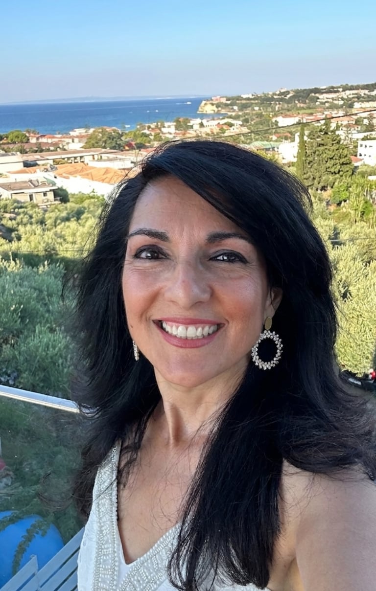 Smiling woman with dark hair on a balcony overlooking a Mediterranean coastal village.