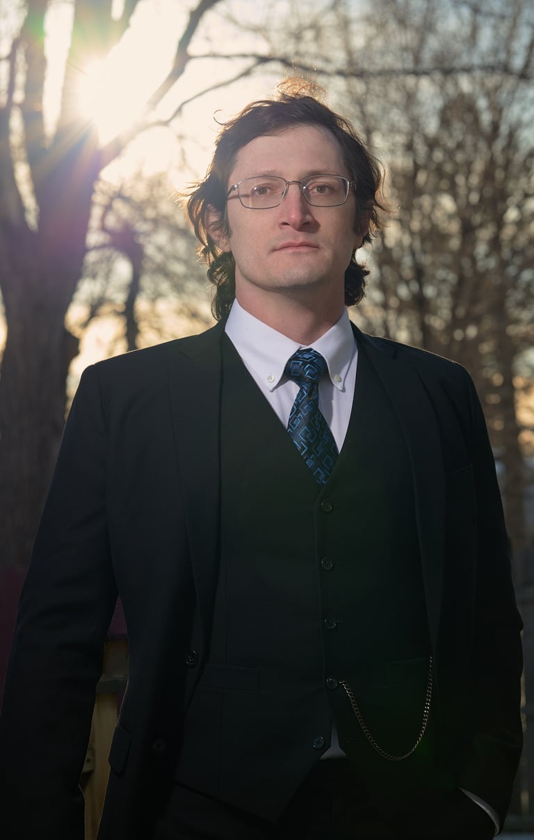 a man in a  black suit and glasses and tie with a blue tie in front of the sun and trees