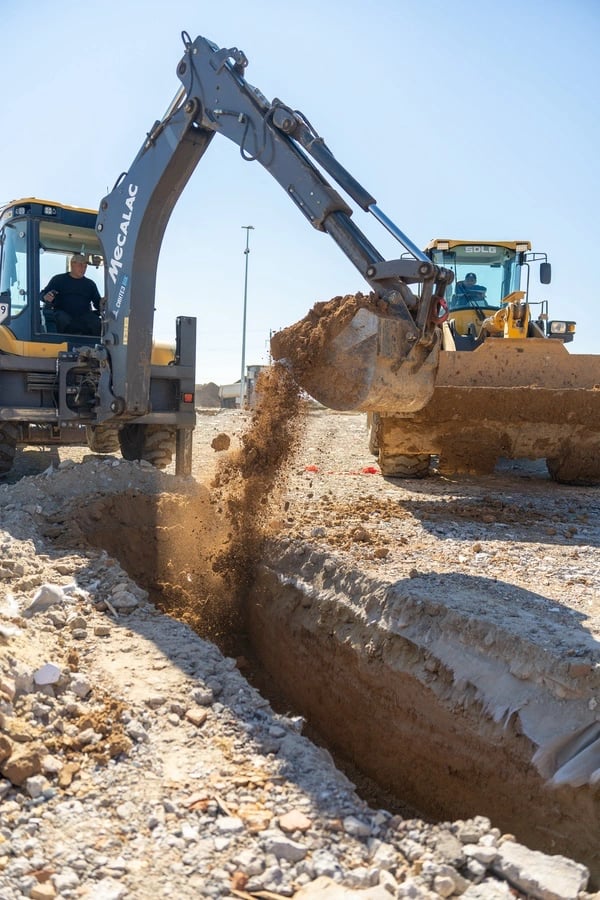 Na zdjęciu widać duży żółty koparkę, która kopie rów na budowie. Koparka ma potężny wysięgnik i szer