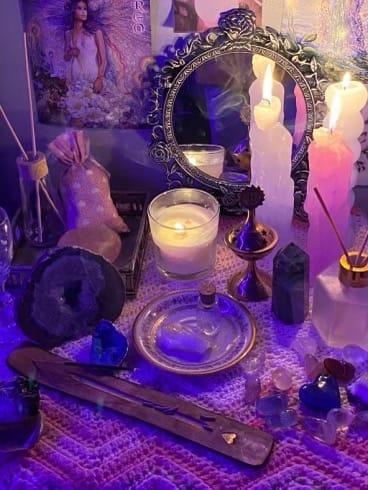 A mystical spiritual altar with lit candles, incense, crystals, and a vintage mirror under purple lighting.