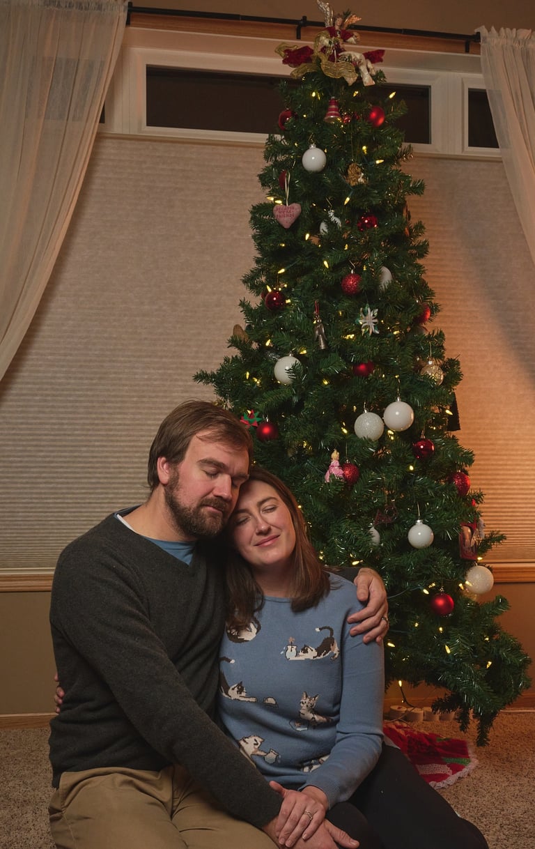 A couple hugging and sitting in front of a decorated Christmas tree with warm lights and ornaments.