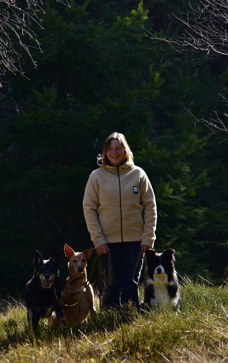 Smiling woman in a fleece jacket standing with three dogs in a sunny evergreen forest.