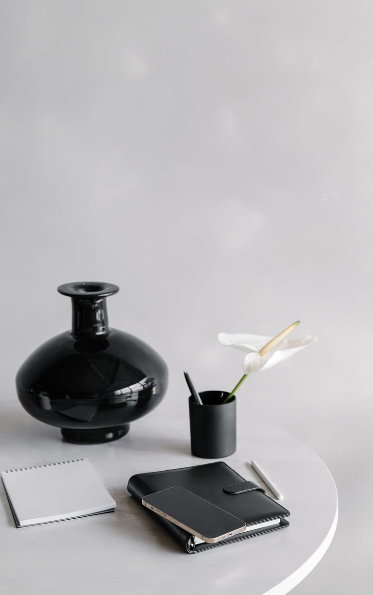 a black vase, a flower in a pencil cup, and notebook on a white desktop