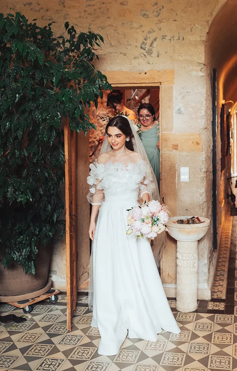 Anaïs, prête pour son mariage, sortant du gîte en robe de mariée avec son bouquet.
