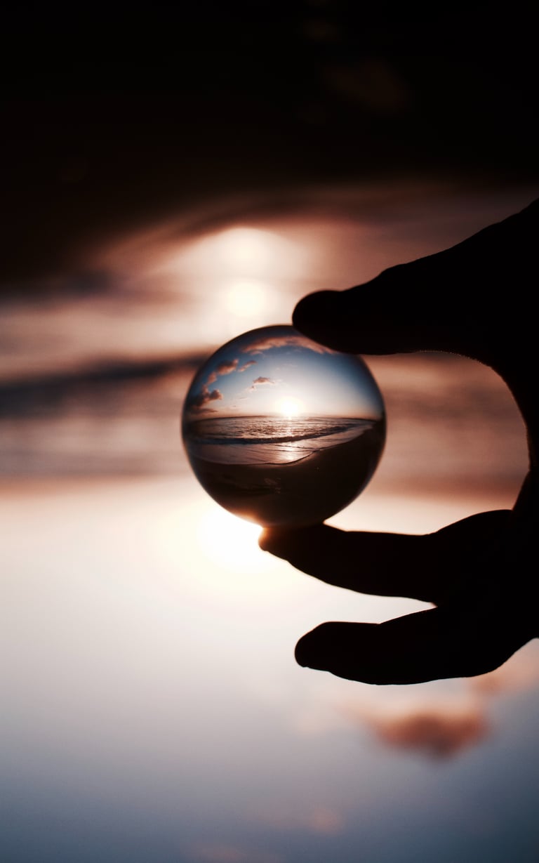 Des doigts tenant une boule de cristal reflétant avec clarté un coucher de soleil sur la mer