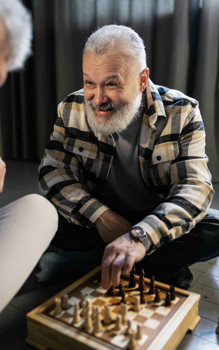 Homem idoso sorridente, com barba branca e camisa xadrez, concentrado em uma partida de xadrez no chão.