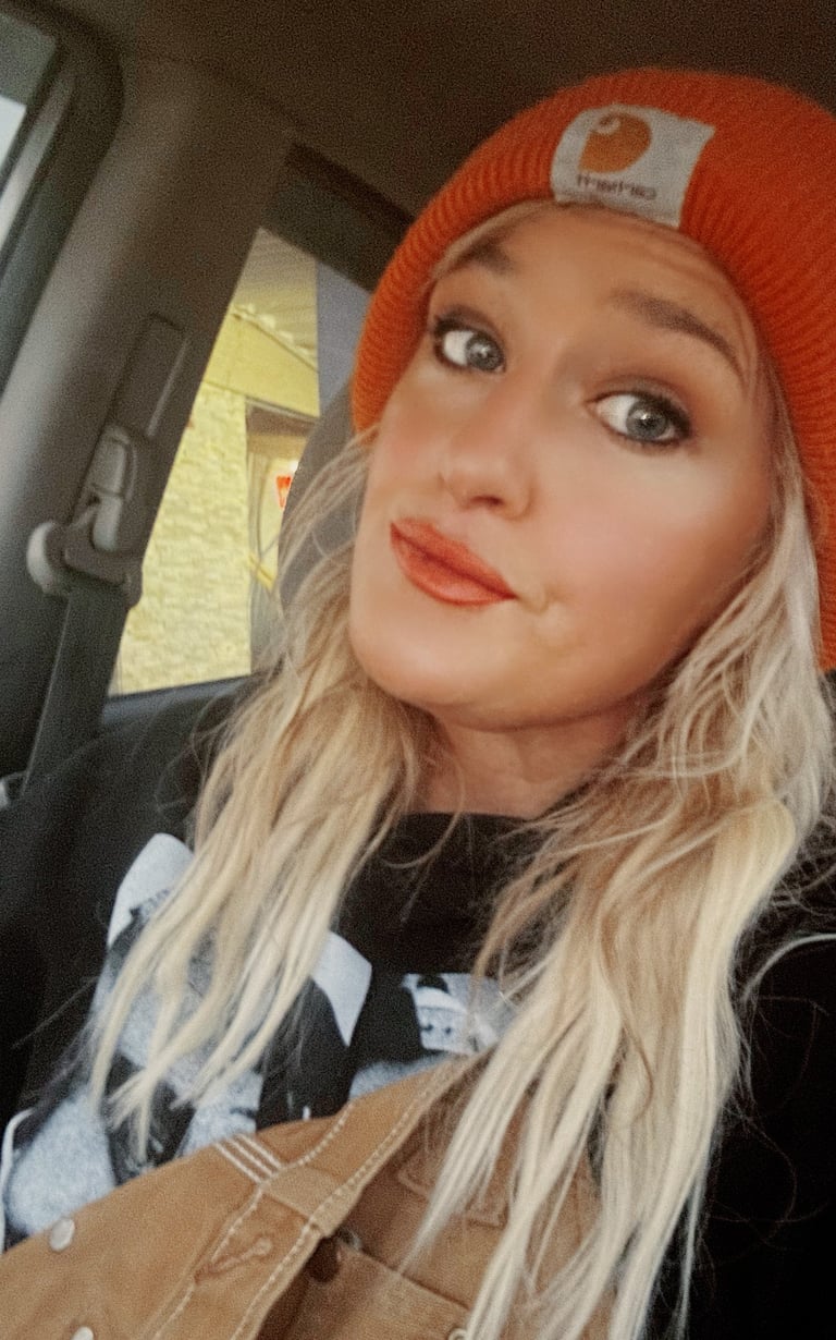 Blonde woman wearing an orange Carhartt beanie and black sweatshirt sitting in a car.