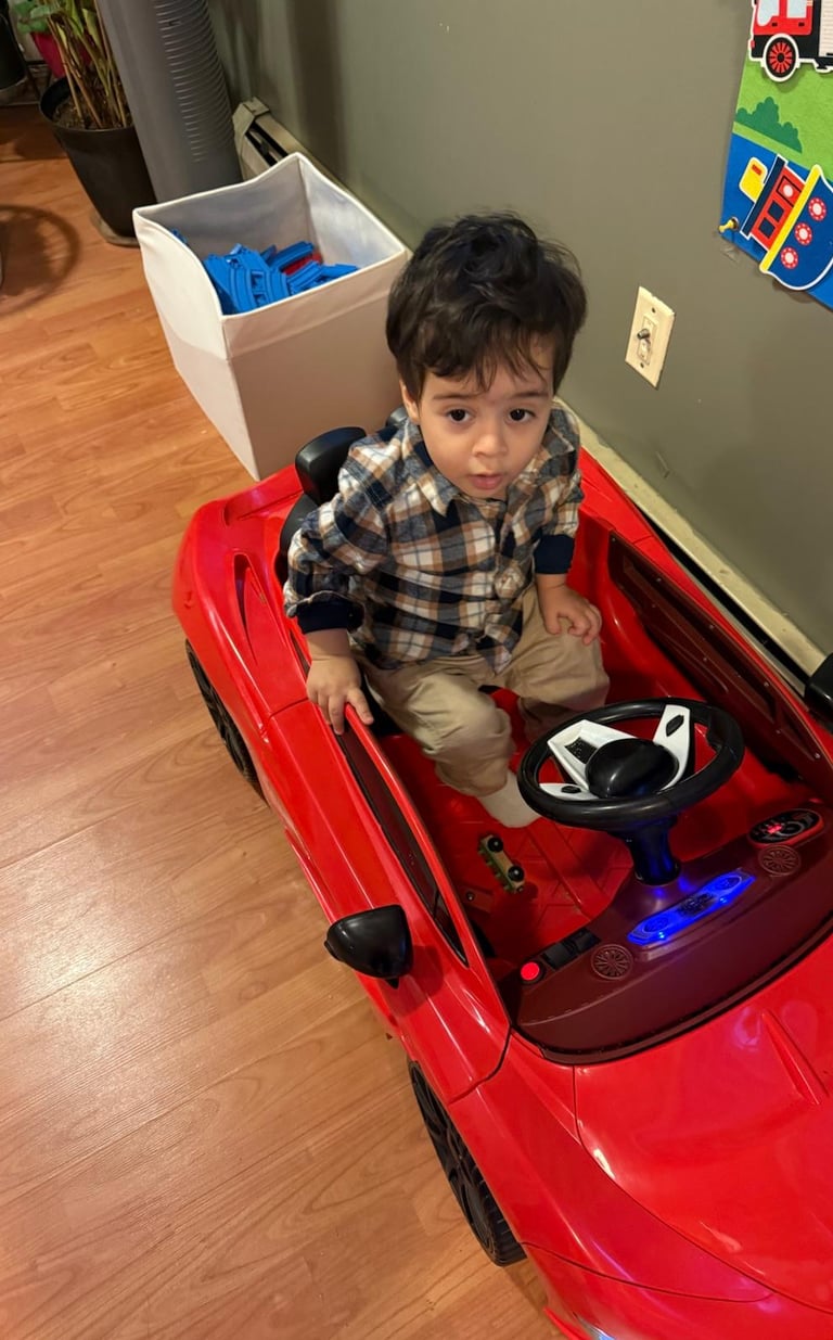 A young boy sitting in a red electric ride-on toy car in a playroom.