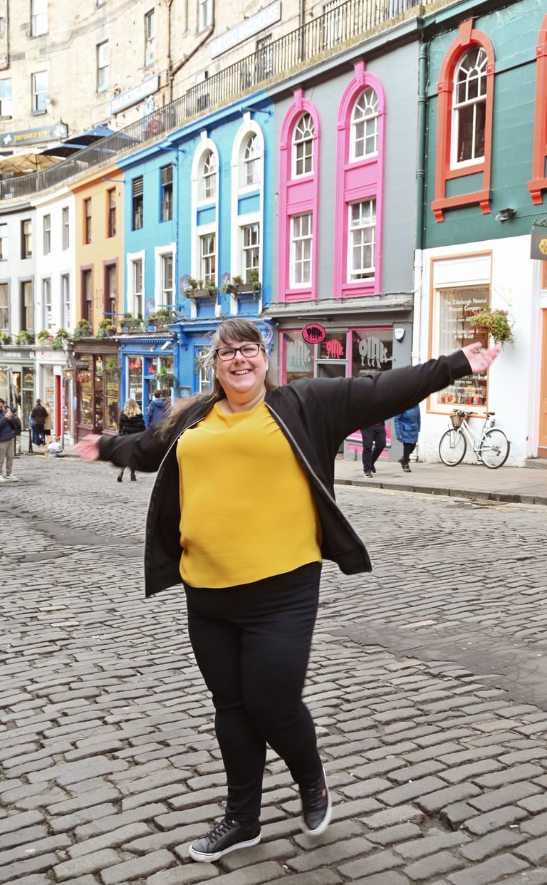 Laura is dancing with her arms out on a cobbled street with brightly coloured buildings behind her