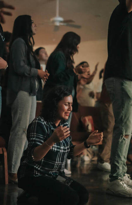 Person kneeling with eyes closed and hands raised, surrounded by others in communal worship.