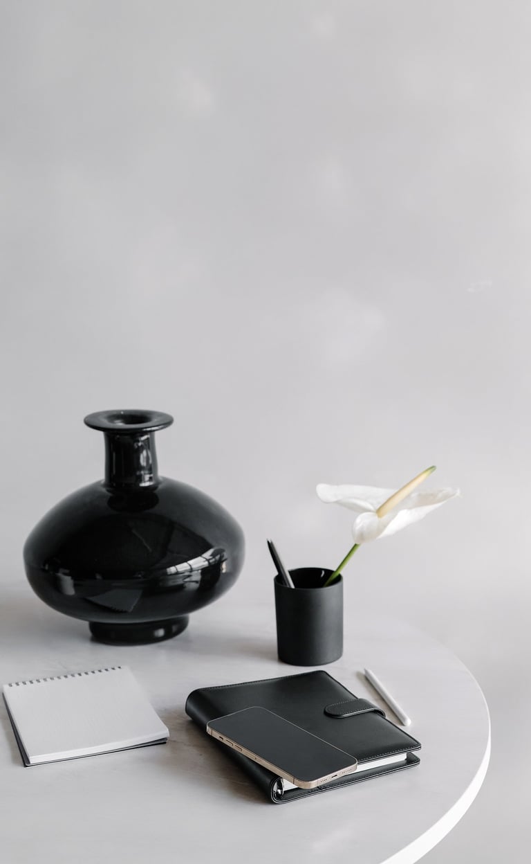 a black vase, a flower in a pencil cup, and notebook on a white desktop