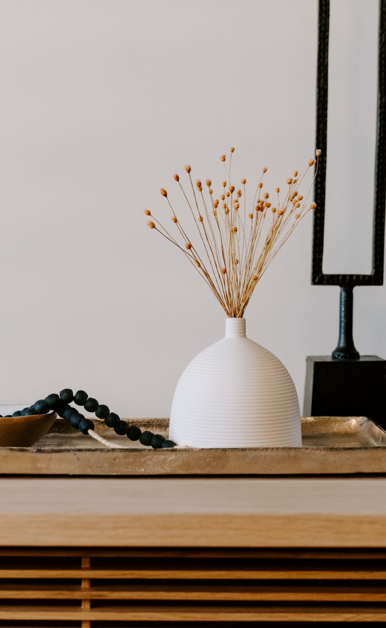 Minimalist home decor featuring a white ribbed vase with dried flowers on a gold tray with black beads.