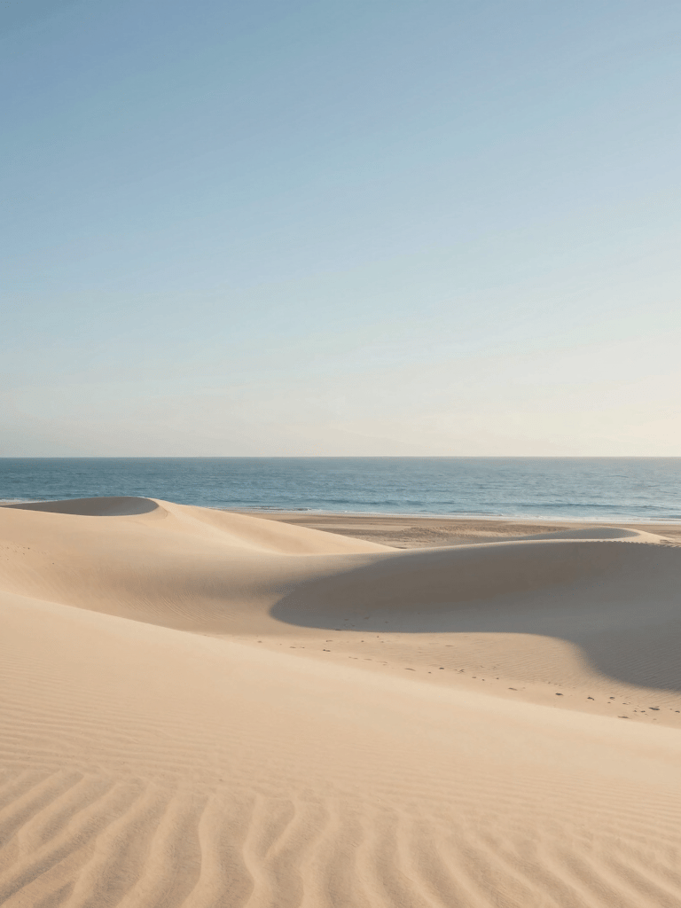 A minimalist fine-art photography of a serene Mediterranean beach in Spain. In the composition, fine sand beige dunes gently meet the calm, translucent sky blue sea. The morning light is soft and warm, casting long delicate shadows on the ivory-colored sand. Clean, spacious layout conveying deep tranquility, peace, and high-end minimalism.