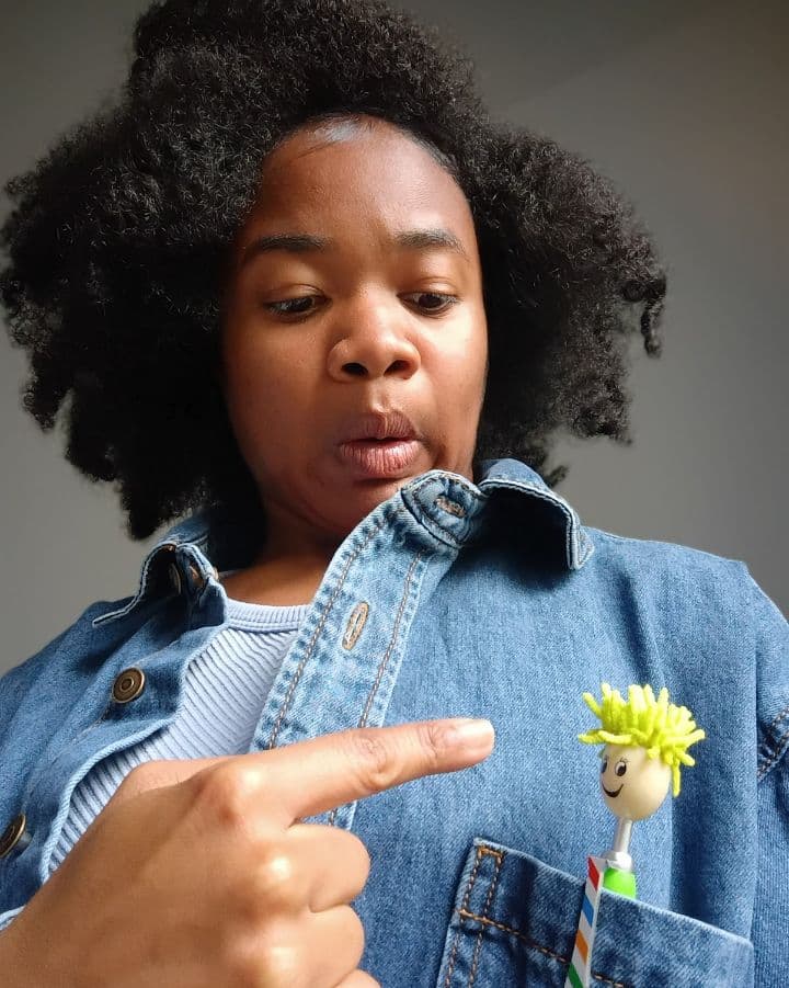 A woman in a denim jacket points at a small smiley face toy in her pocket with a surprised expression.
