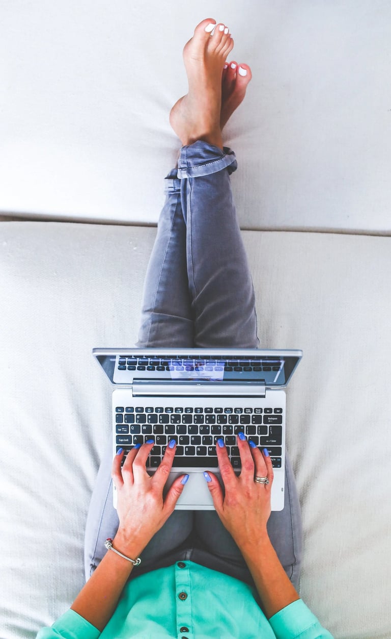 Femme en train de pianoter sur son ordinateur portable et qui a les jambes allongées sur le canapé blanc