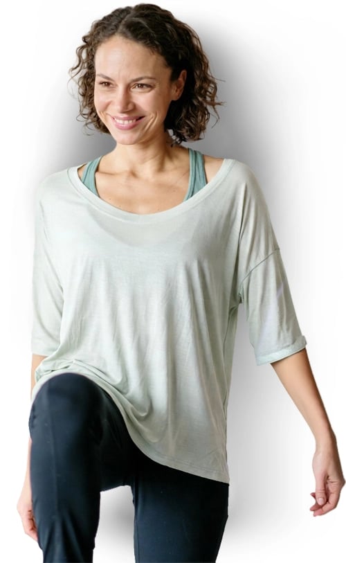 A smiling woman wearing a loose light green yoga top and black leggings performs a fitness stretch.